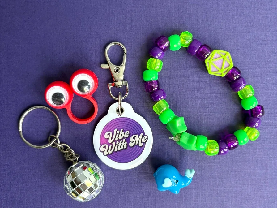 A Vibe Tag wearable NFC keychain with the Vibe With Me logo, shown alongside a kandi bracelet, disco ball keychain, and rave accessories